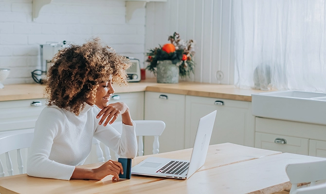 African American woman smiles holding credit card in front of open laptop sitting at table in farmhouse kitchen.