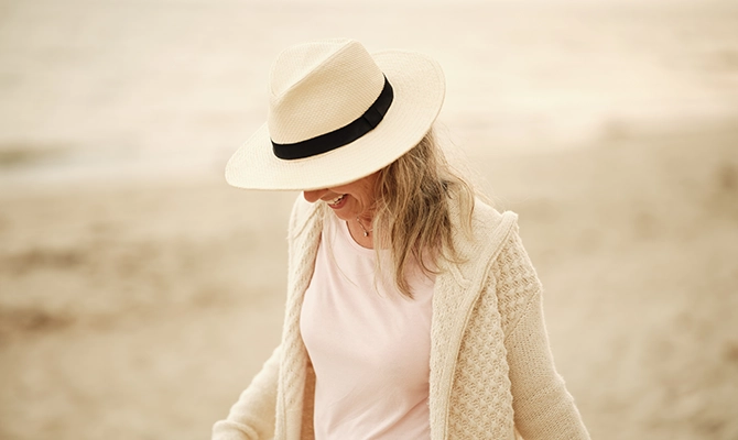 Older woman walks Harwich beach head down looking at the sand, smiling with a hat on just covering her face