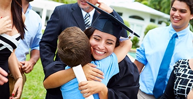 College graudate in cap and gown holding diploma hugs little brother while smiling.