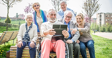 Group of six older adults sitting on park bench together smiling and viewing screen of an ipad