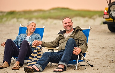 Older husband and wife sit on beach chairs holding hands at Sandy Neck Beach.