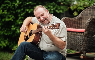 Older man sitting on patio outside playing his guitar and smiling.