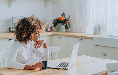 African American woman smiles holding credit card in front of open laptop sitting at table in farmhouse kitchen.