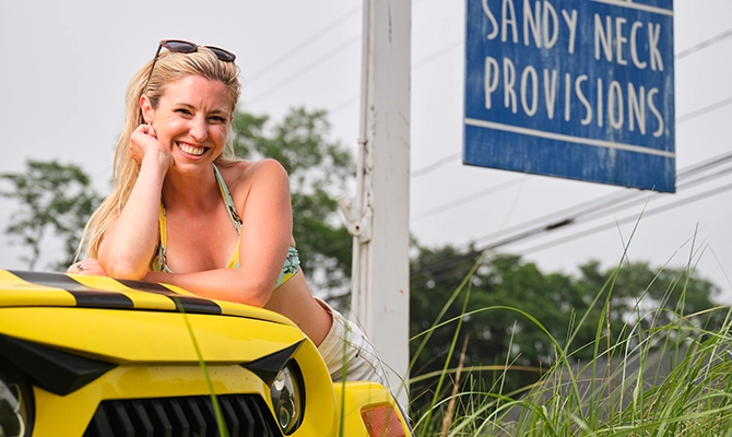 Local Cape Codder and owner of Sandy Neck Provisions, poses proudly on her yellow Jeep in front of her storefront.