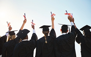 Graduation, back of students dressed in cap and gowns, arms up holding diplomas, celebrating